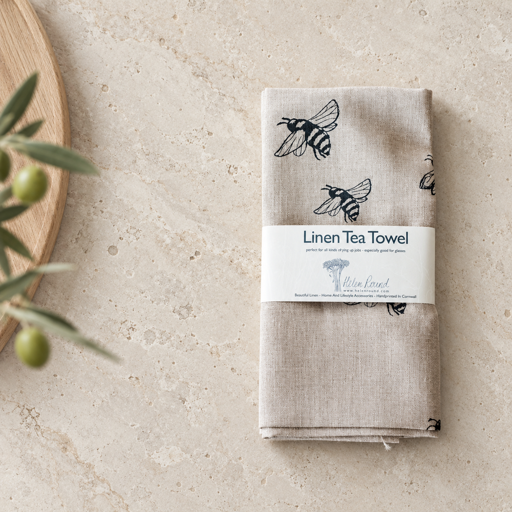 Folded Helen Round natural linen tea towel with black bee illustrations and paper packaging on a pale stone worktop beside an oak board and olive sprig.