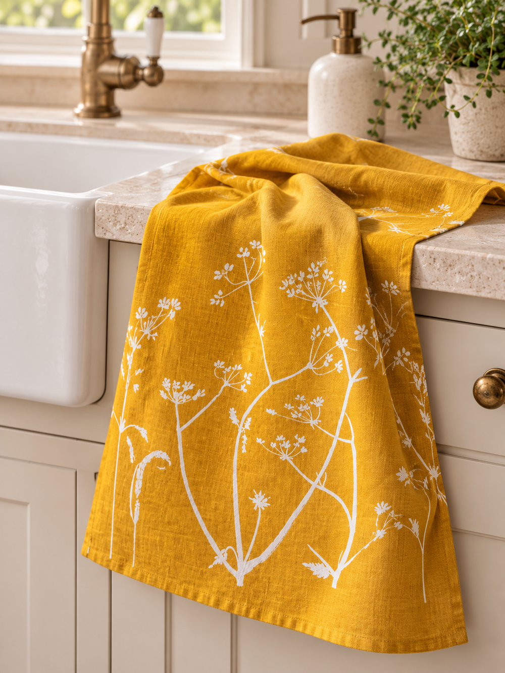 Mustard linen tea towel with a white hedgerow design draped beside a farmhouse sink in a cream kitchen with brass fittings.