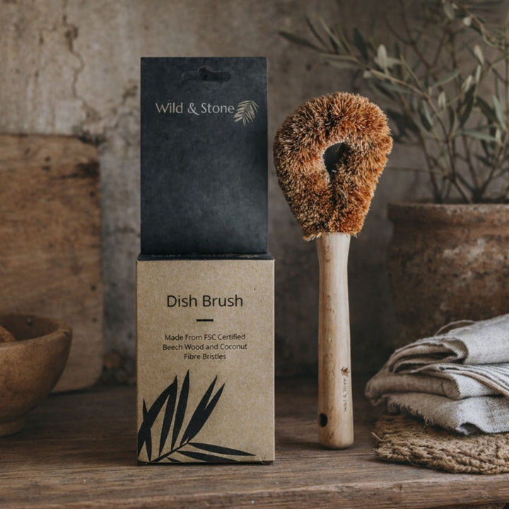 Wild &amp; Stone coconut fibre dish brush with FSC beech wood handle beside eco packaging on rustic kitchen surface