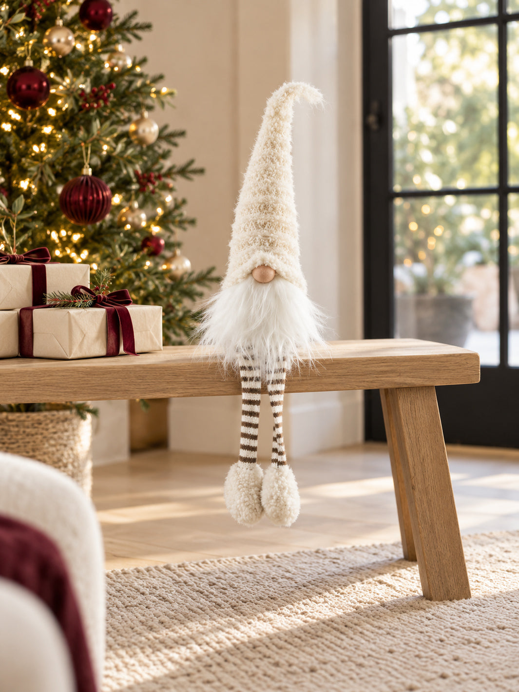 Cream Christmas gonk with a tall fluffy hat, white beard and brown and white striped legs sitting on an oak bench beside a decorated Christmas tree.
