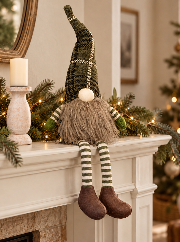 A Christmas gnome decoration hanging from a white fireplace mantel in a cozy living room. The gnome has a tall dark green knit hat with white stripes, a shaggy brown beard, green and white striped sleeves, and brown boots.