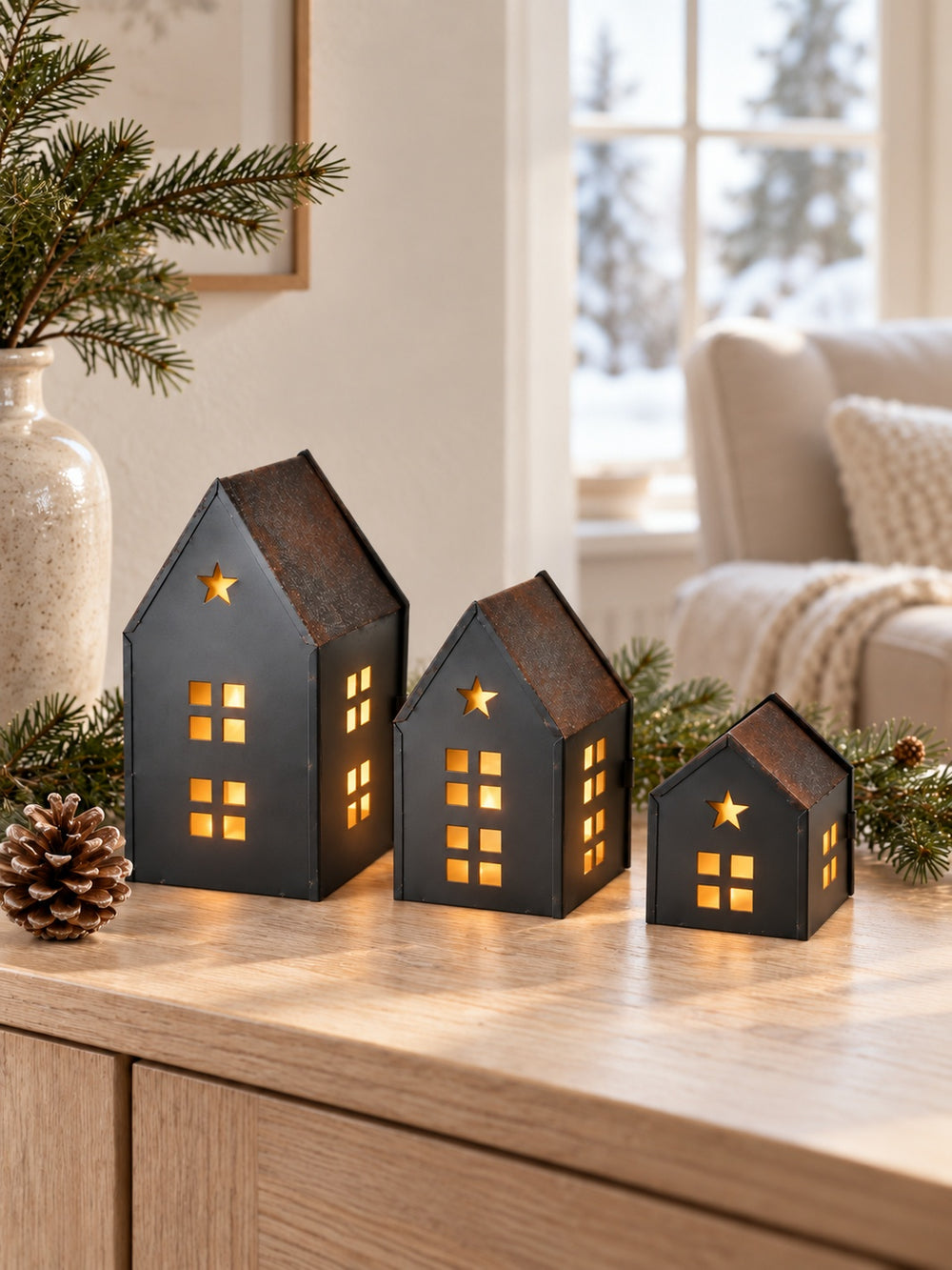 Three illuminated charcoal metal houses with rustic brown roofs on an oak console beside pinecones and winter greenery.